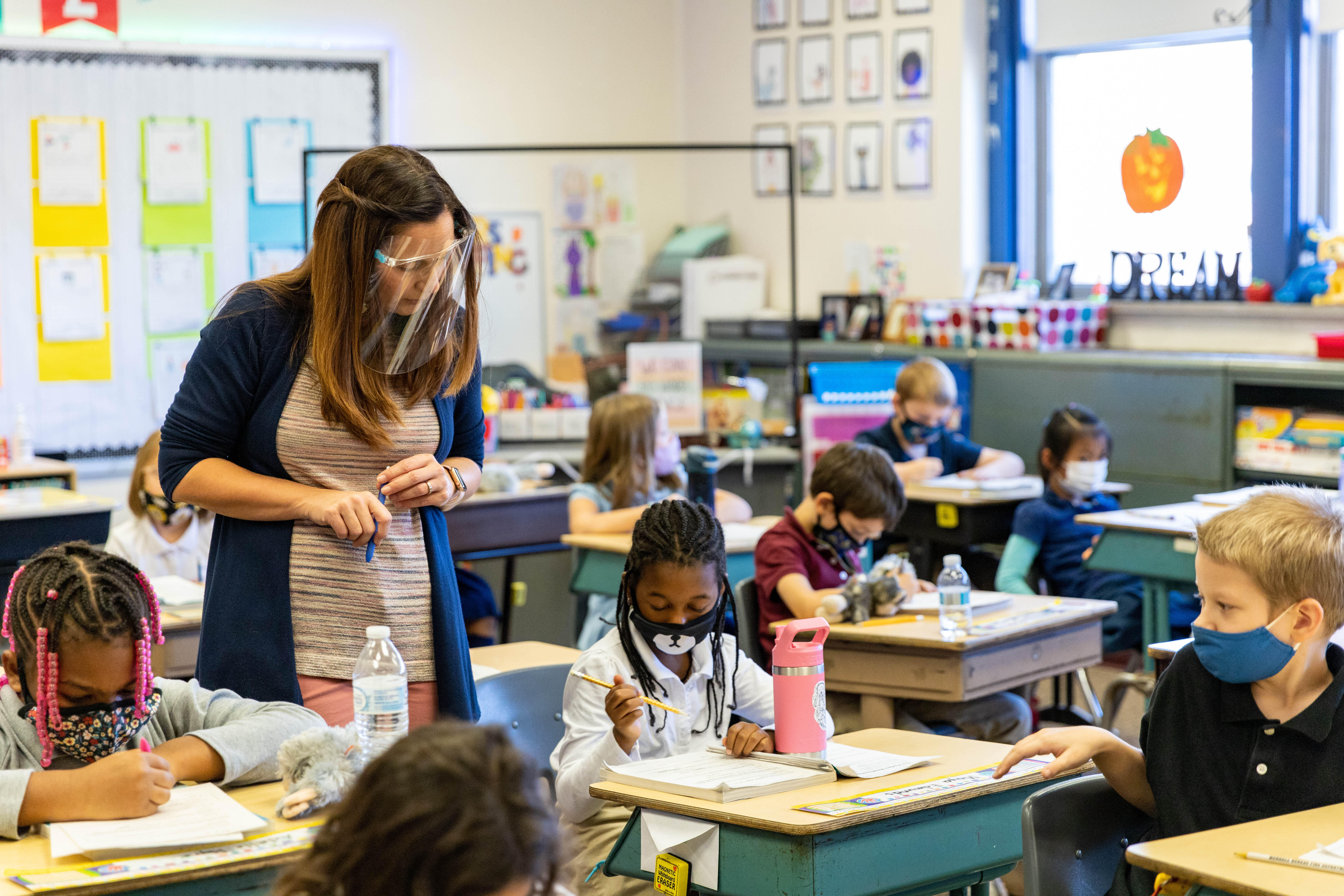 A teacher watches over an elementary-aged student as she works in a workbook. 