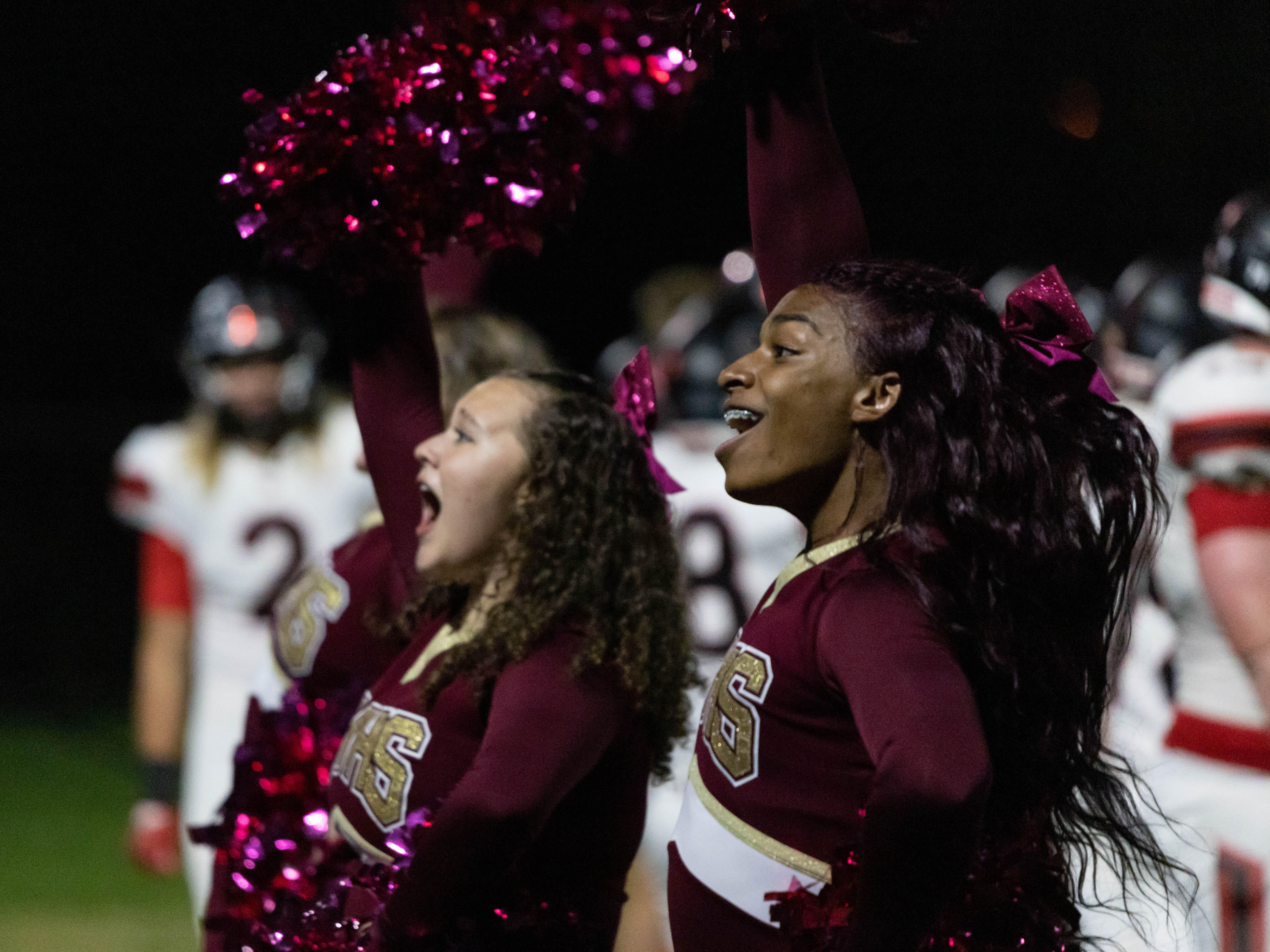 Two cheerleaders raise their pompoms in the air and yell out during a high school football game.