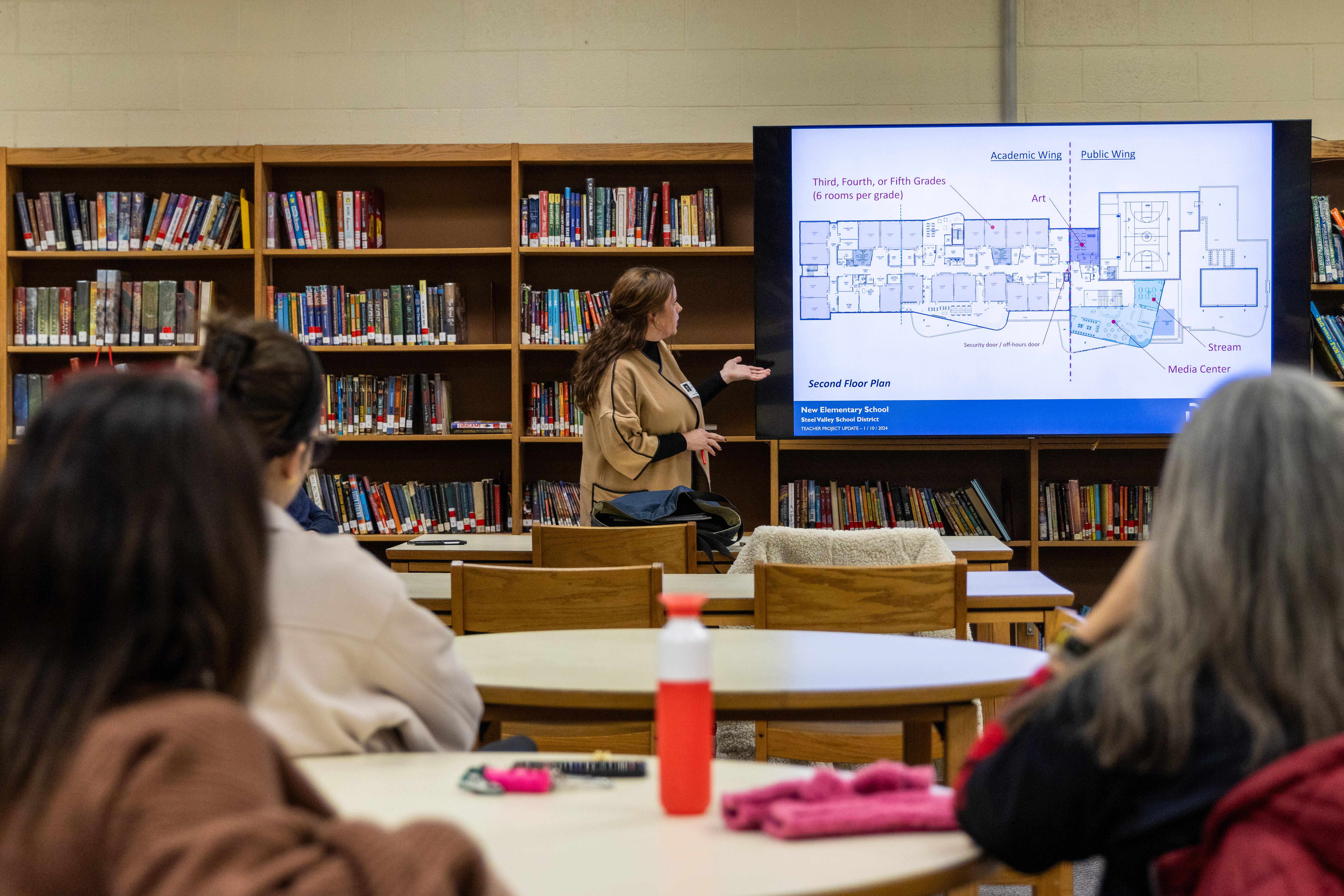 A woman stands at a digital screen and gestures to a blueprint of a building while talking to an audience in a school library