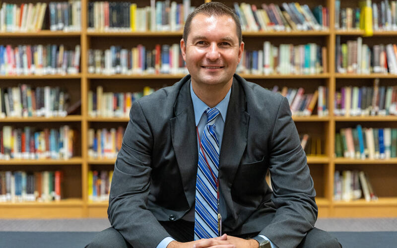 Assistant superintendent Bryan Macuga sits in front of a bookshelf in the library at Steel Valley High School.