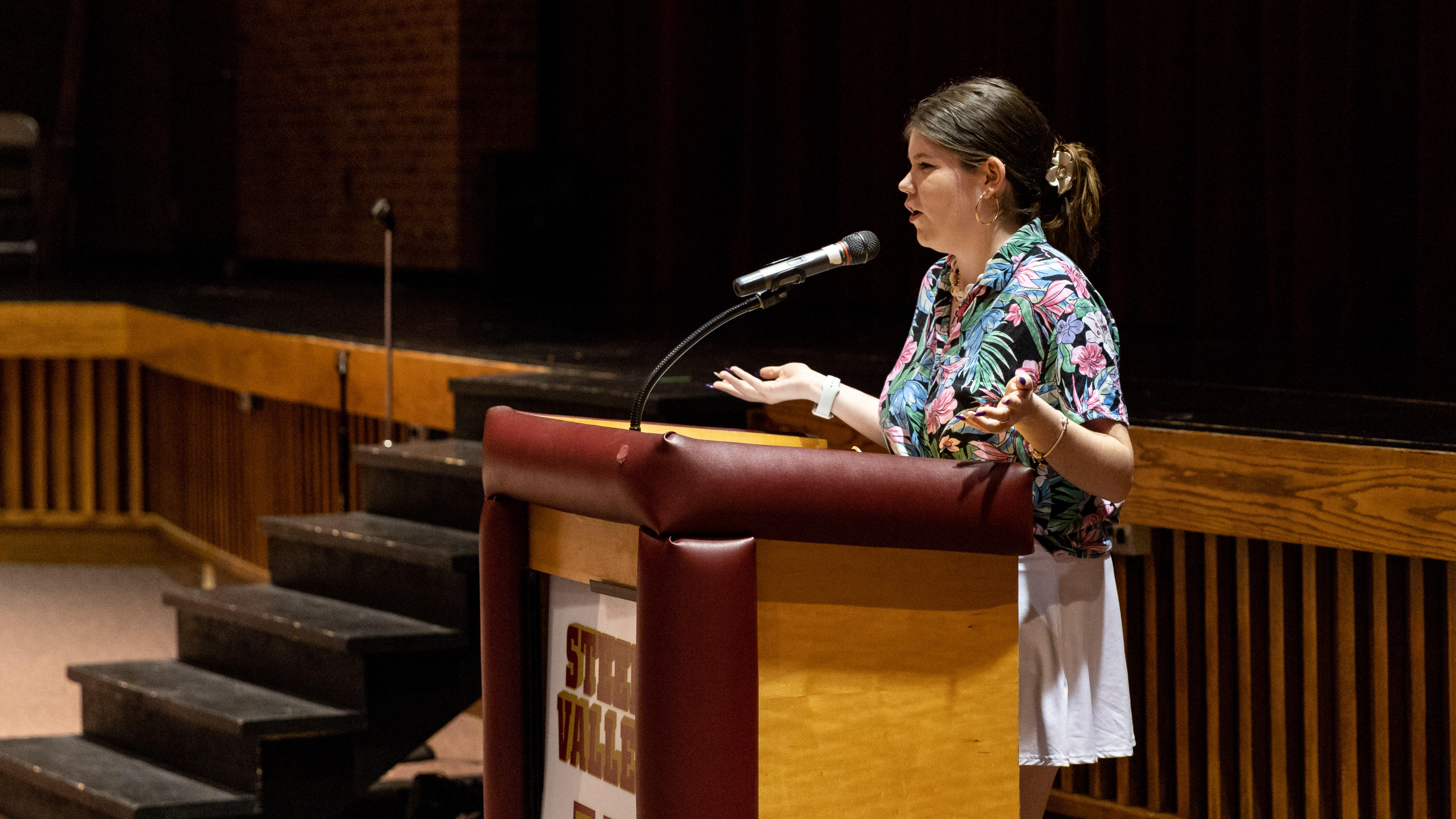 A young woman stands at a podium in an auditorium and addresses the seated audience.
