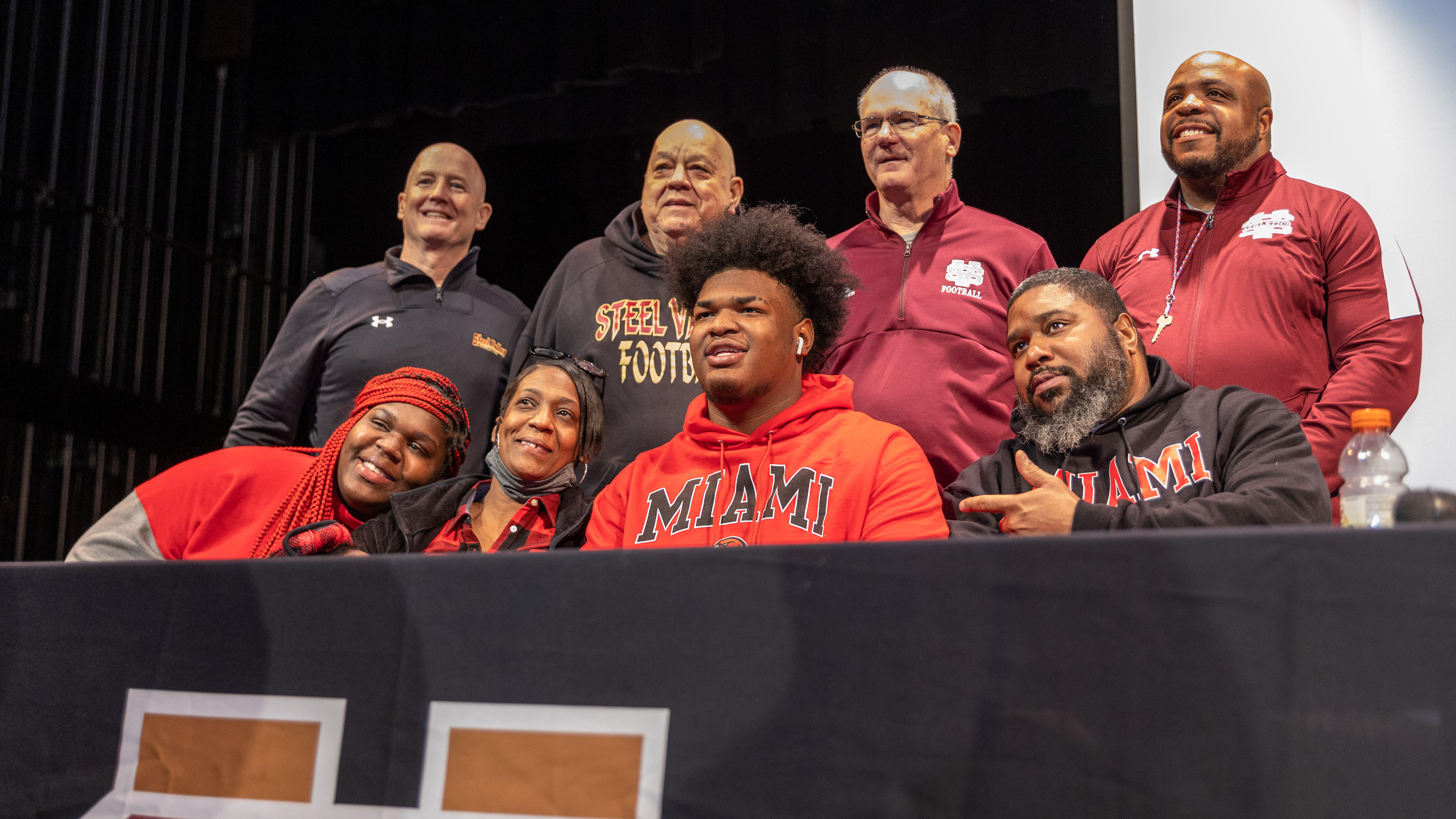 Greg Smith poses for a photo with his family and coaches. He is seated at a table on a stage.