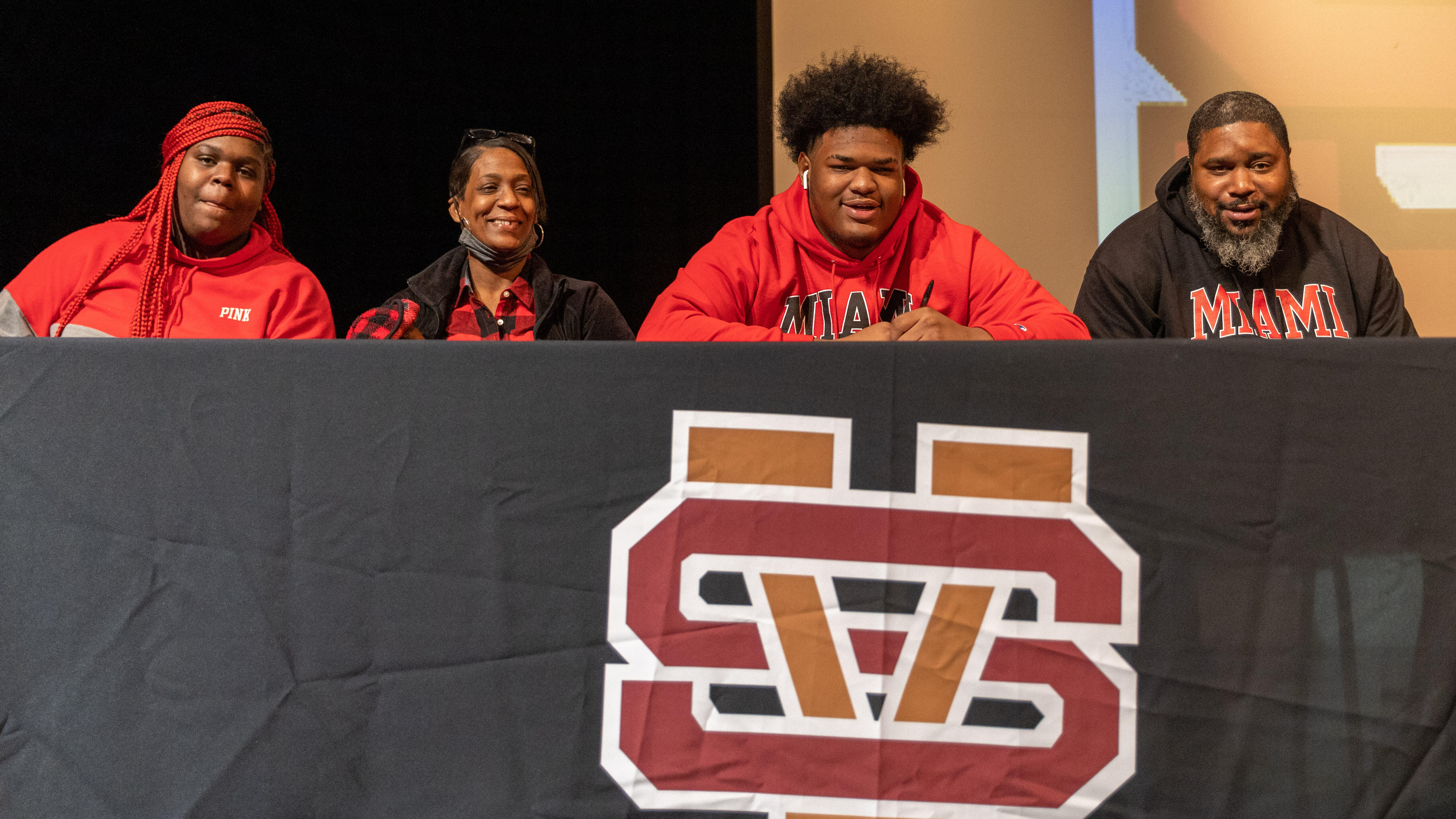 Greg Smith signs his National Letter of Intent while surrounded by his family. He is seated at a table on a stage.