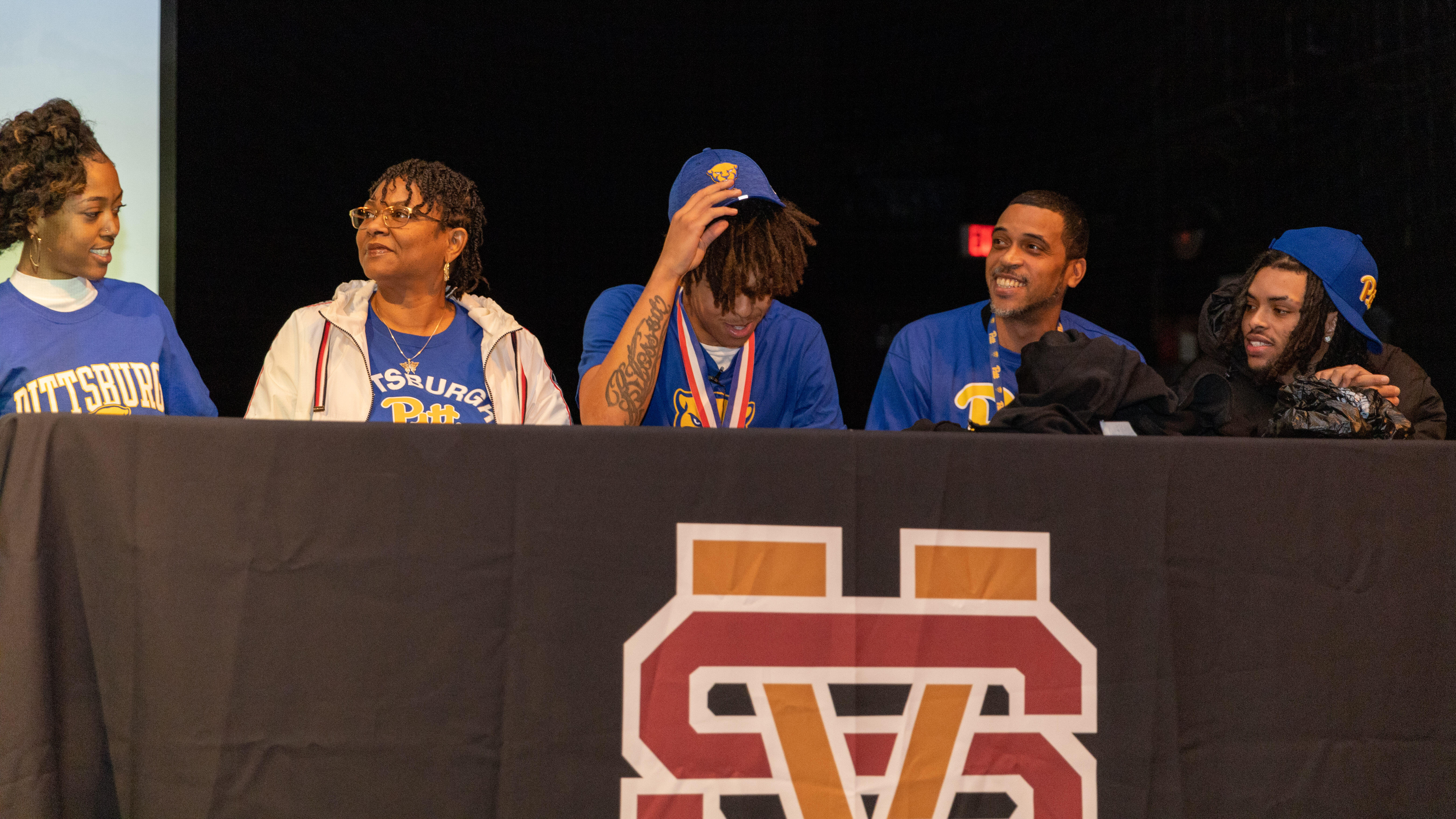 Cruce Brookins reveals his Pitt shirt underneath a hoodie to announce where he's going to college. He is seated at a table on a stage.