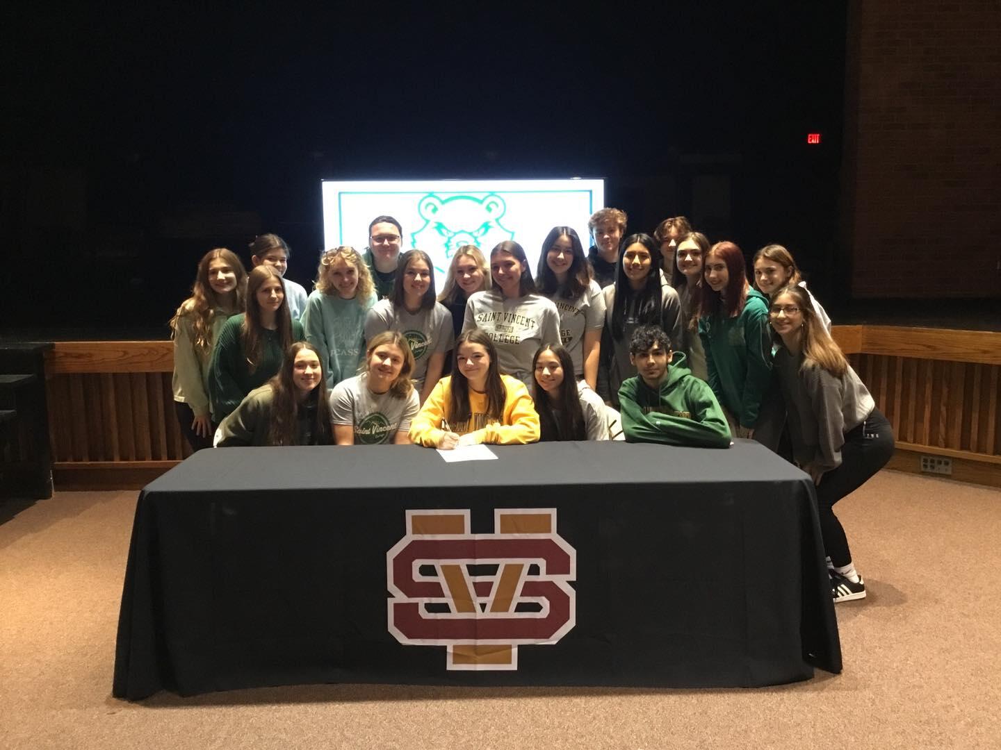 A student-athlete poses with her teammates behind a table. She is seated while they stand around her as she signs a document.
