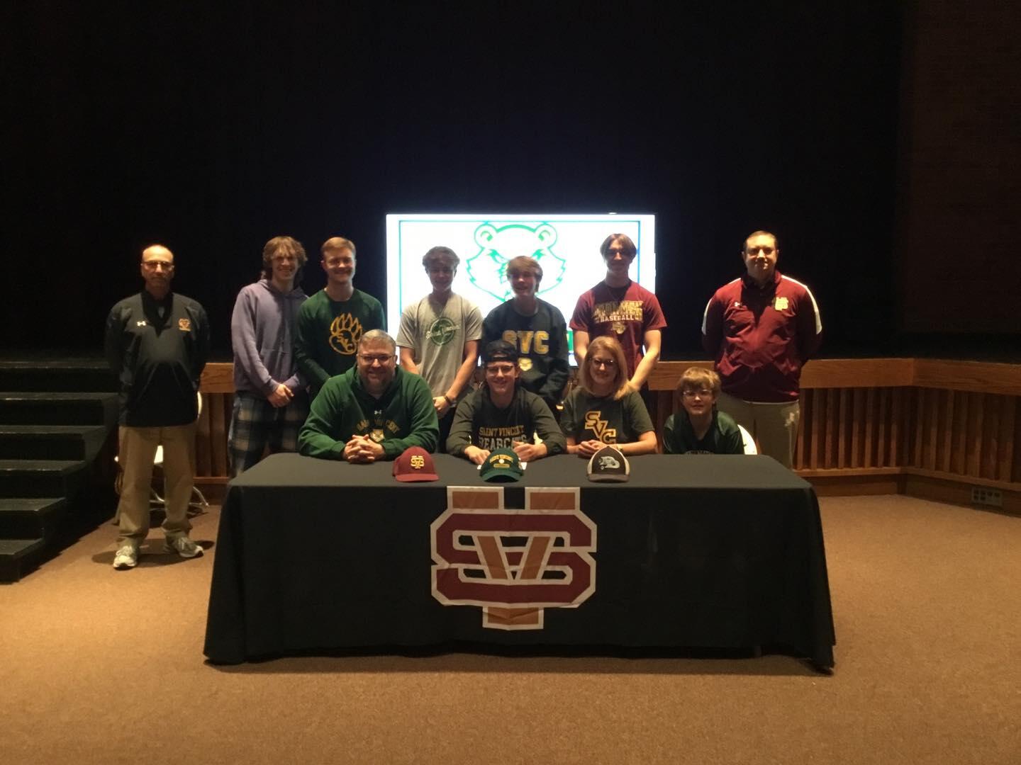 A student-athlete poses with his teammates behind a table. He is seated while they stand around him as he signs a document.