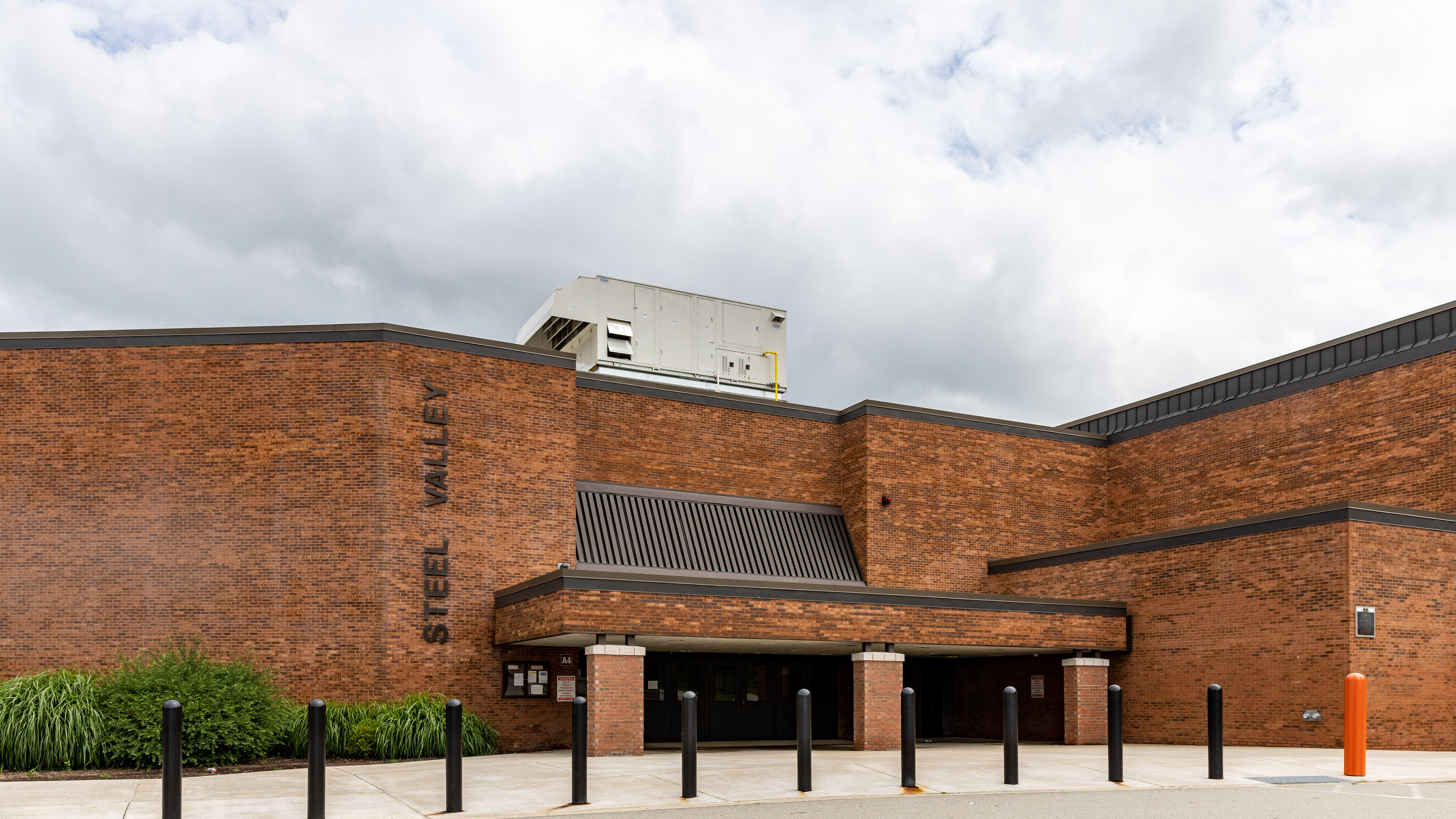 An image of the Steel Valley High School entrance on a cloudy day. It is an orange-brown brick two-story multi-story building with a covered entrance supported by columns.