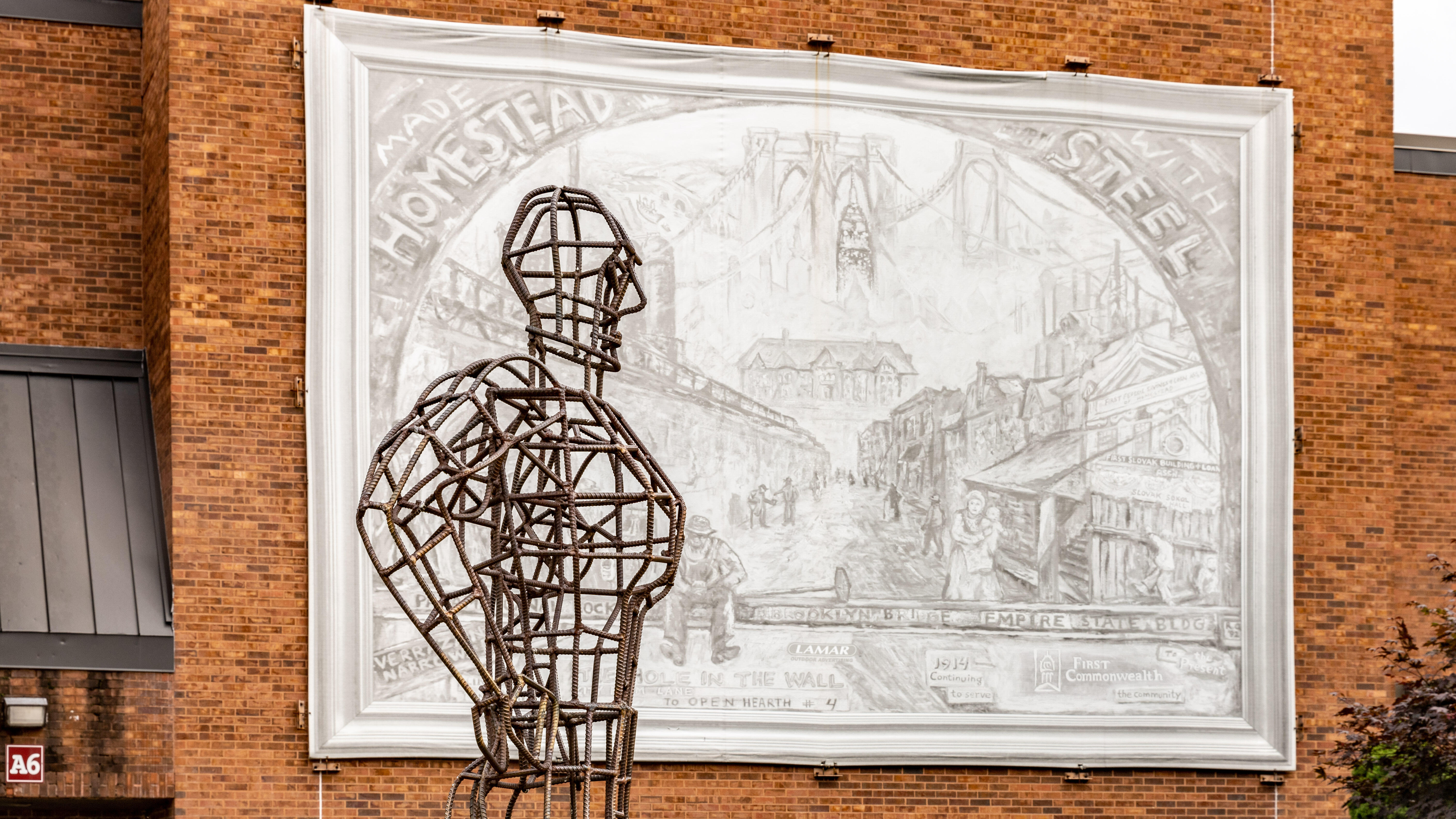 A wire frame sculpture of a person stands in front of an engraved relief on the side of the Steel Valley Middle School/High School building. 