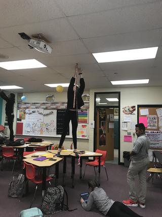 Student stands on desk and holds measuring stick to ceiling