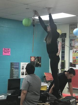 Student stands on desk and drops doll and stick