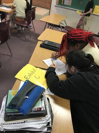 Two students work on paper next to folder marked Exhibit C