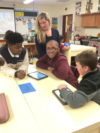 One student smiles at camera while others look at tablets on table