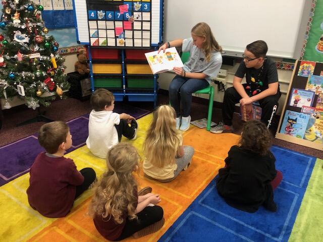 Two middle school students read to a group of younger students. The young students are seated on colorful mats on the floor while the older students are in chairs. 